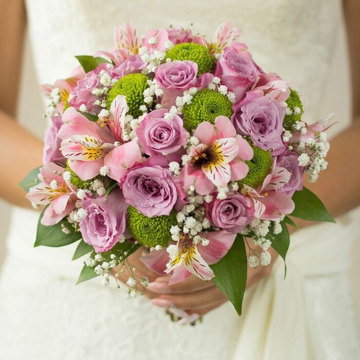 Showcasing the creativity of South London flower artistry, this luminous bridal bouquet is tenderly held by a bride against the soft texture of an ivory lace gown. The masterful bouquet unfolds in a harmonious sphere, bringing together layers of pastel lavender and blush pink roses, each dewy petal alive with subtle gradients of colour and light. Between these classic blooms, spherical lime green button chrysanthemums pop with modern flair, their fresh palette evoking the bright spirit of Peckham's community gardens and the lively shades found in local markets. Interspersed throughout the arrangement are captivating pink alstroemeria lilies, trumpet-like in form, streaked with maroon and gently dusted with yellow-a nod to the borough's diversity and charm. Wisps of white gypsophila, or baby's breath, create airy frameworks that soften the transitions between each flower, adding a cloud-like lightness that seems to float above the bride's hands. A ring glimmers subtly as fingers encircle the bouquet stems, and a few glossy, deep green leaves complete the lush frame. The close-up scene reveals both the tactile richness of velvet roses and the crisp vibrancy of the mums, a combination that mirrors Peckham's blend of the traditional and the contemporary. This arrangement, expertly designed by Florist Peckham, perfectly exemplifies wedding floral trends and provides local inspiration for couples celebrating love in the heart of SE15.